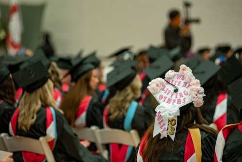 Students sit for graduation ceremony. One grad cap is decorated in white and pink saying 