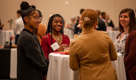 Students stand around a table and smile while listening to a woman who's back is to the 
camera.