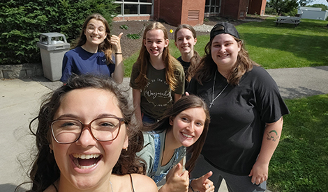 6 young woman stand in front of Garlock on campus and smile while taking a selfie.