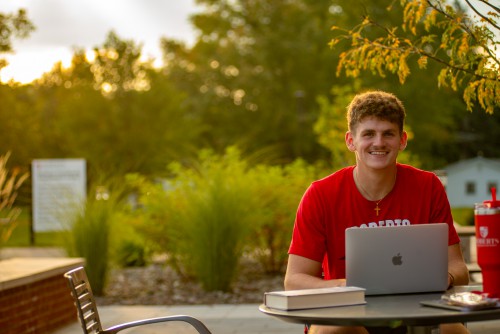 A young male student sits outside in front of a laptop and smiles.