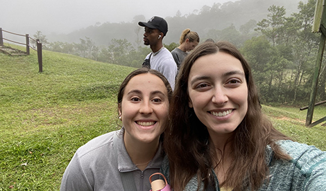 2 young women take a selfie, smiling while the Brazilian sky and fields are in the background.