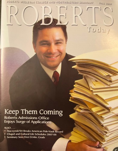 Kirk smiles with a big pile of files as featured on the copy of Roberts Today Magazine