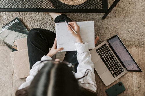 An overhead view of a woman holding a planner and pen. Surrounding her are a laptop and books.