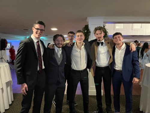 5 young men wear suits and smile with their arms over each other's shoulders. Another young man poses in the background.