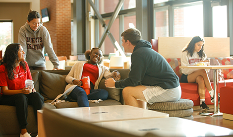 4 students sit, laugh, and talk in the Golisano Community Engagement Center. Another student is seen studying in the background.