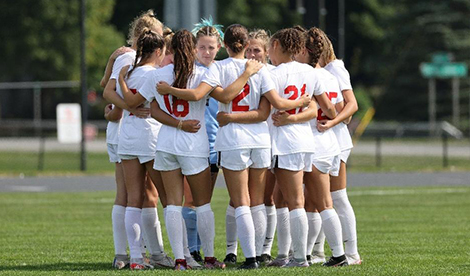 The women's soccer team wears white uniforms and gathers in a circle with their arms around each other.