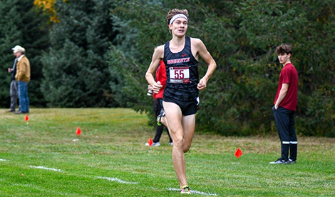 A young man wears a Roberts uniform and runs along the grass in a cross country race.