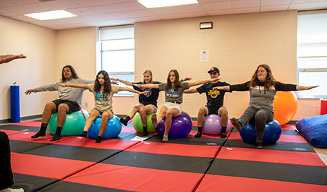 Summer campers sit on exercise balls with arms outstretched.