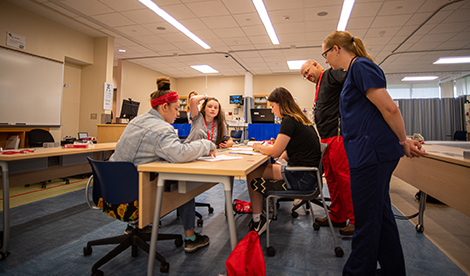 Summer campers do work around a table as 2 professors stand beside the table.