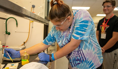 A student does a science experiment. Another student is seen in the background smiling.