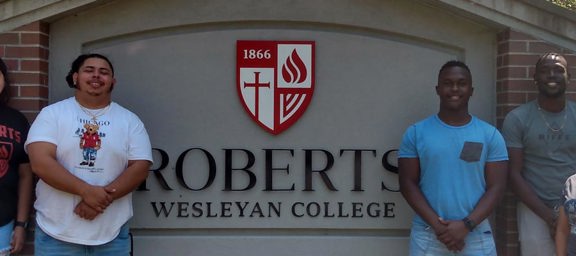 6 students smile in front of a sign saying Roberts Wesleyan College