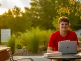 A young man sits with a laptop open and smiles